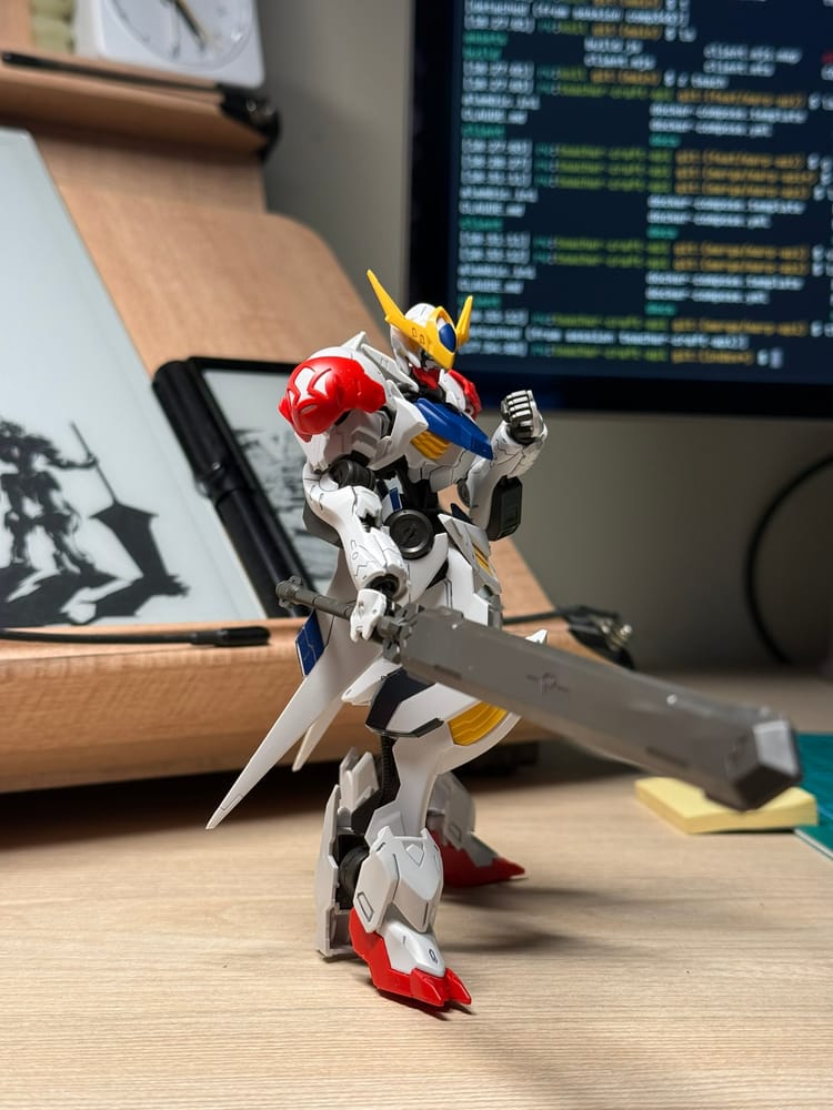 A white, red, and blue Barbatos model holding a large sword on a desk in front of a code editor