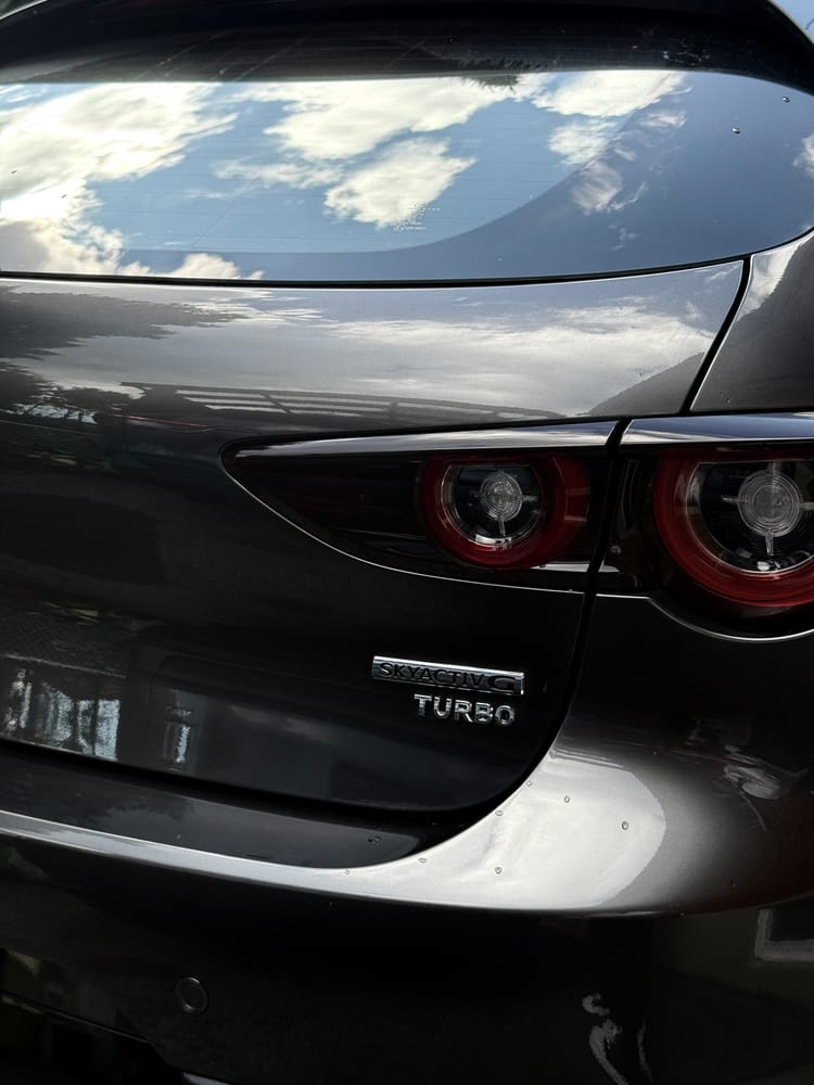 The rear of a dark Mazda with round taillights, a turbo badge, and clouds reflected in the glass