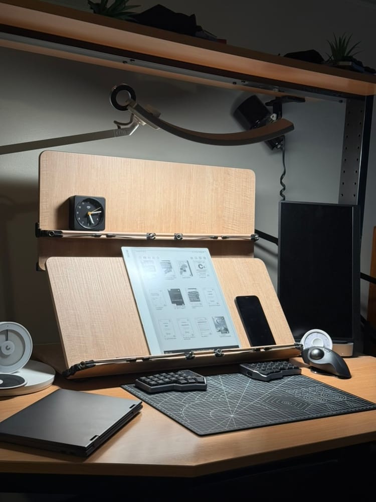 A wooden reading stand holding an e-ink tablet and phone above a split keyboard and cutting mat