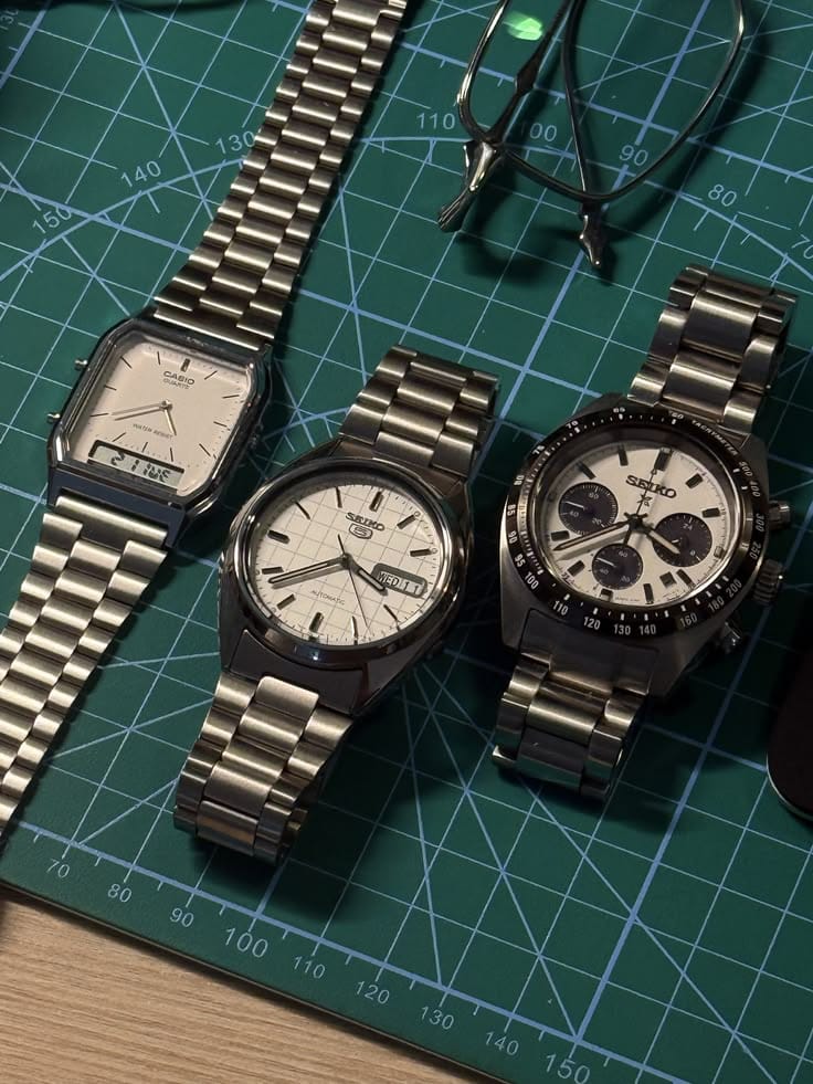 Three silver Casio and Seiko watches beside glasses on a green cutting mat